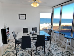 A dining area with a glass table and chairs, a ceiling fan, a bookshelf, and a large window with a view of the ocean.