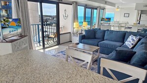 Un salon moderne doté d’une grande fenêtre, d’un canapé bleu, d’une table basse et d’un téléviseur diffusant une scène de plage tropicale.