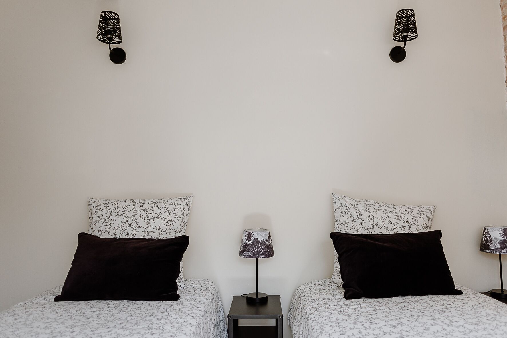 A bedroom with two beds, each with a floral-patterned bedspread and a dark pillow. There are two wall-mounted lamps with a decorative design.