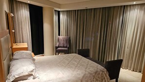 Ein modernes Hotelzimmer mit einem großen Bett, einem Sessel, einem Schreibtisch und einem großen Fenster mit Vorhängen.