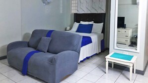 A hotel room with a grey sofa, a bed with blue and white bedding, a small white table, and a large mirror. - Beachfront Escape (Ocho Rios)
