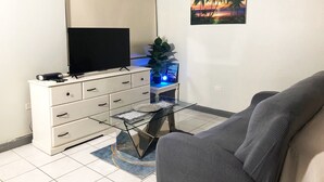 A living room with a flat-screen TV on a white cabinet, a glass coffee table, a gray sofa, and a painting of a sunset on the wall. - Beachfront Escape (Ocho Rios)