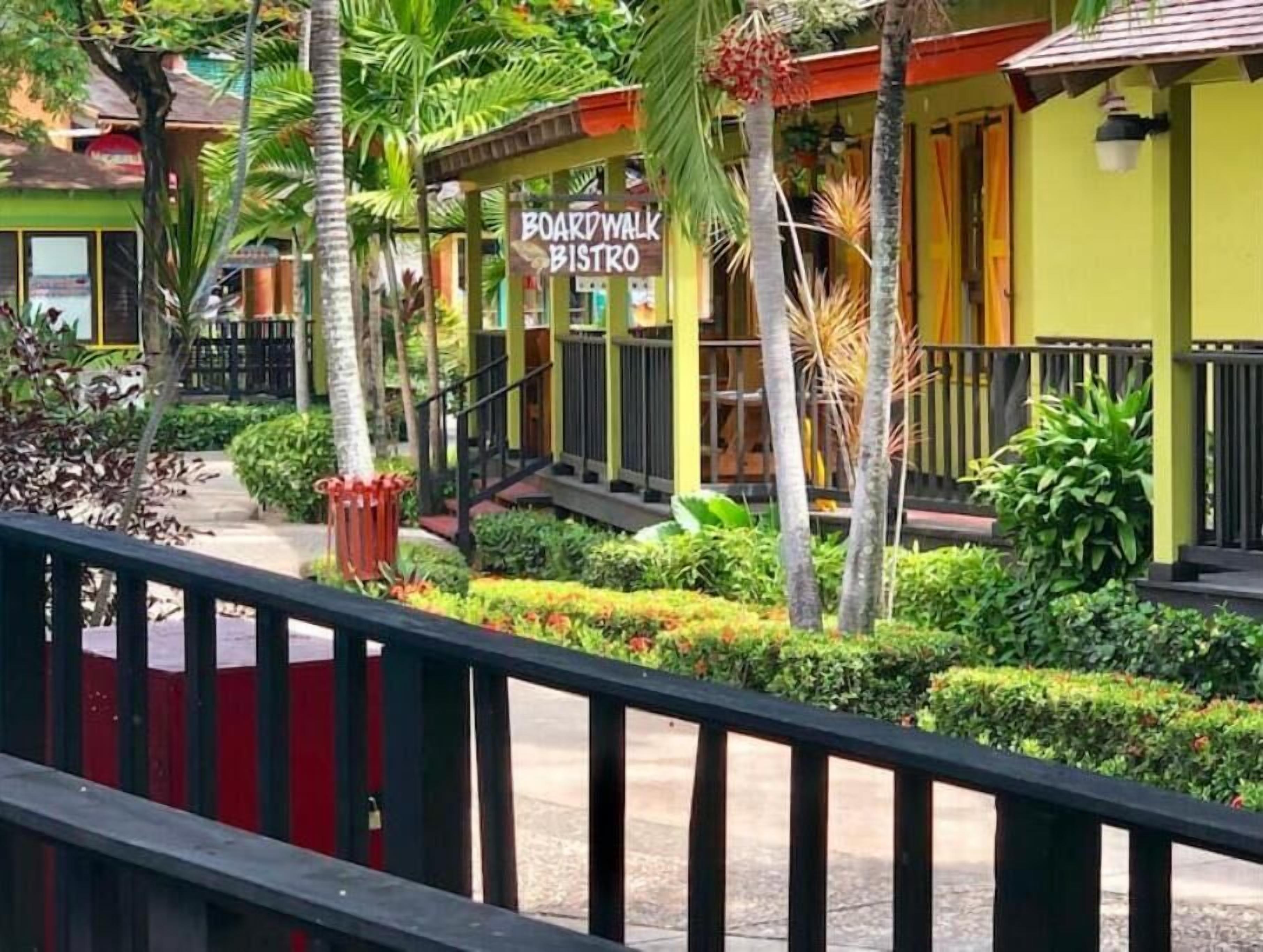 Un bistrot situé sur une promenade en bois, avec une façade verte, des palmiers et une rambarde rouge.