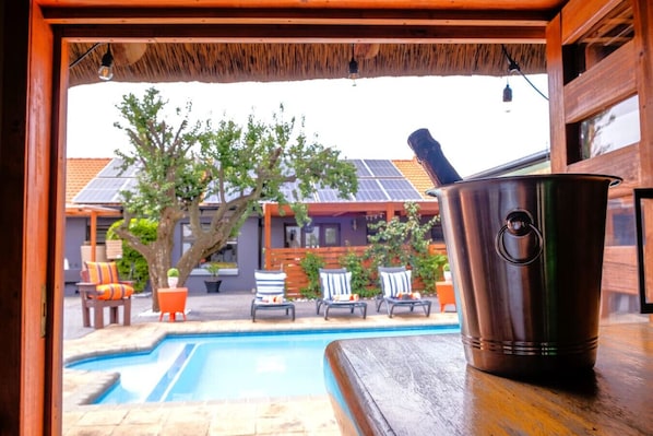 A metal bucket on a wooden table, a swimming pool, and outdoor seating with chairs and potted plants.