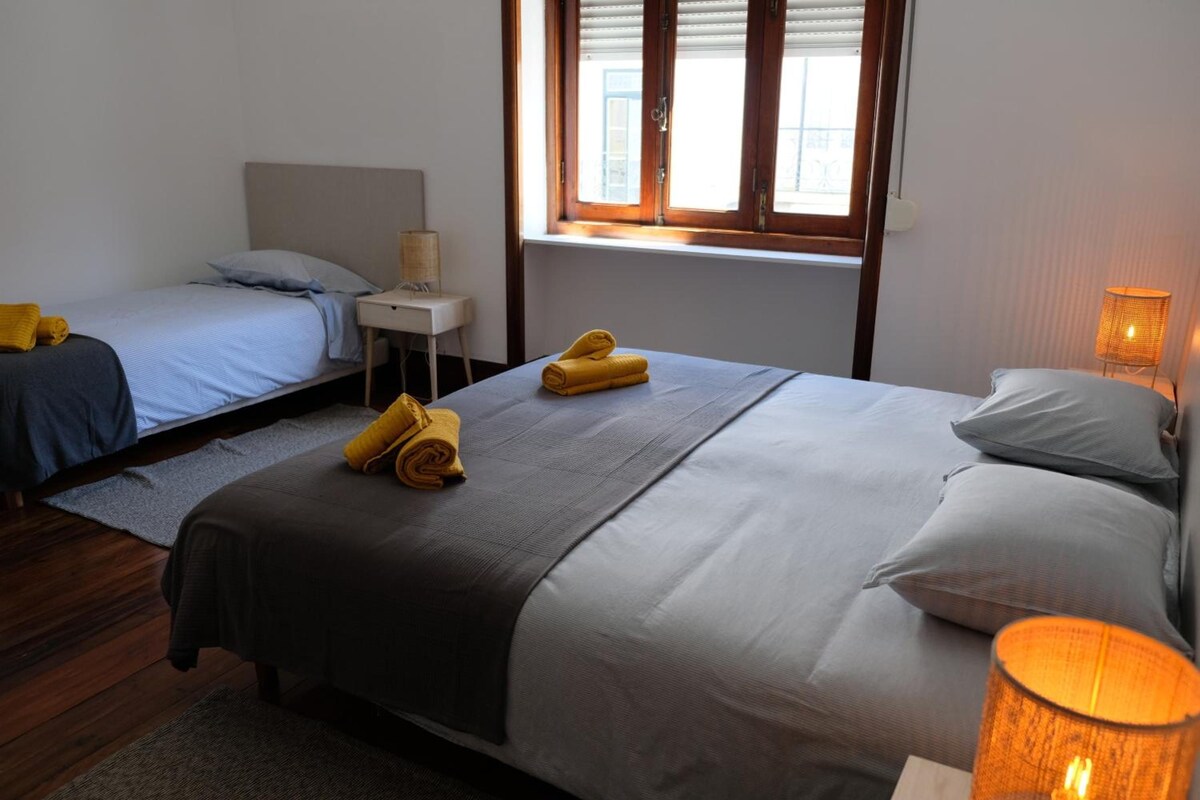 A hotel room with two beds, a nightstand, a lamp, and a window with blinds.