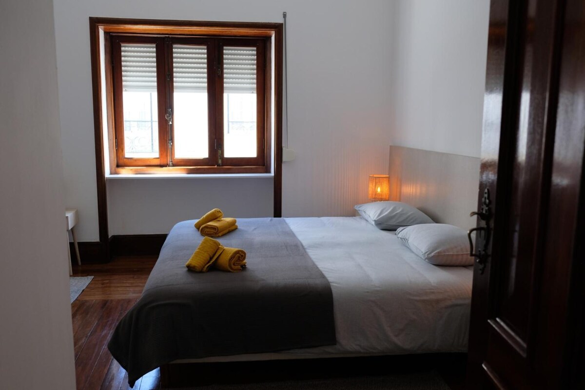 Una cama individual con edredón gris y blanco, dos toallas enrolladas, una lámpara de noche y una ventana con persianas.