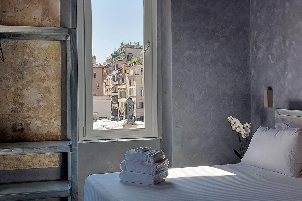 A hotel room with a bed, a window with a view of buildings, a stack of towels, and a vase with flowers.