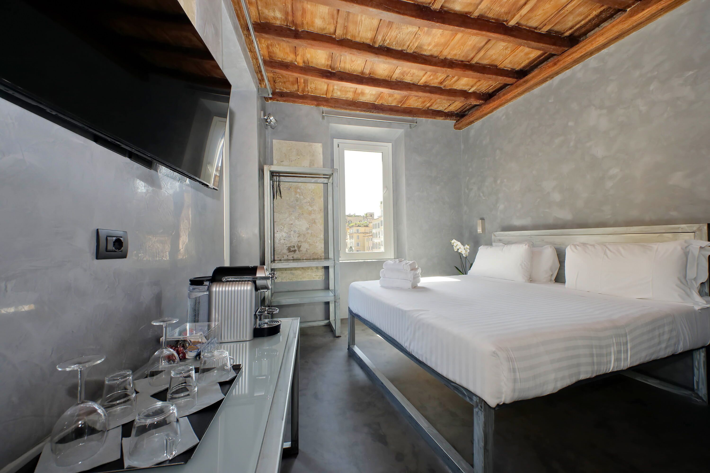 Une chambre d’hôtel avec un lit, une table avec des verres et une bouilloire, une fenêtre et une vue sur les bâtiments environnants.