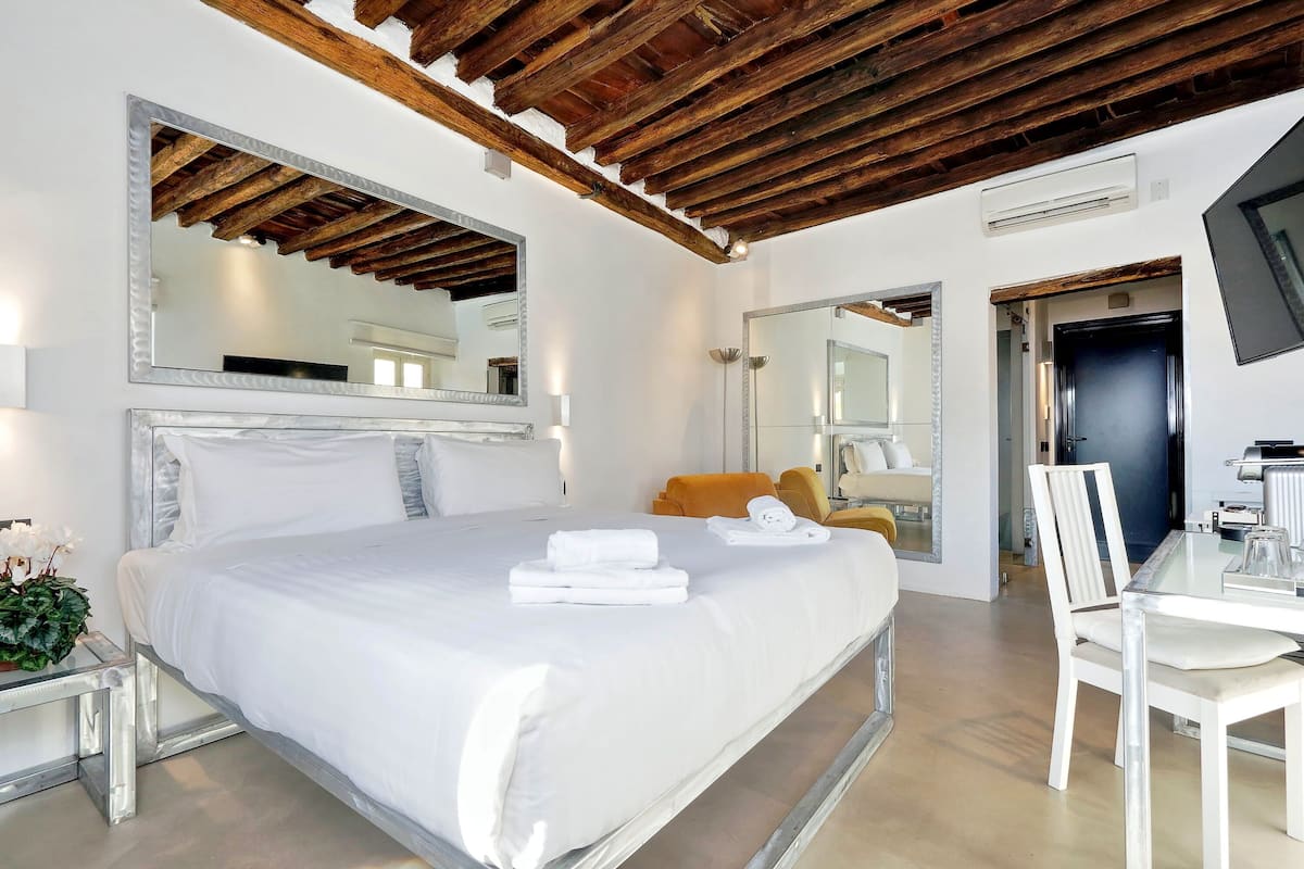 A modern hotel room with a large bed, a wooden ceiling, a flat-screen TV, and a dining area with a table and chairs.