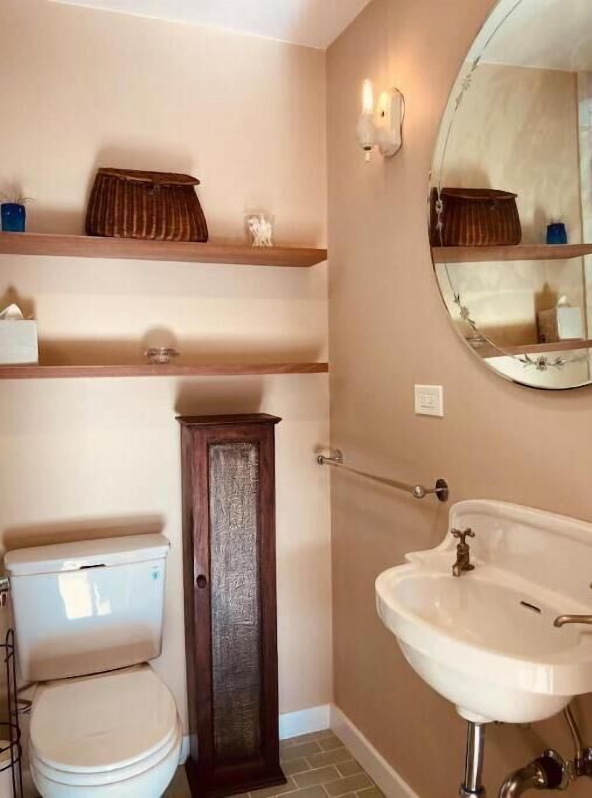 A bathroom with a toilet, a sink, and a mirror. There are shelves above the toilet and a cabinet next to it.