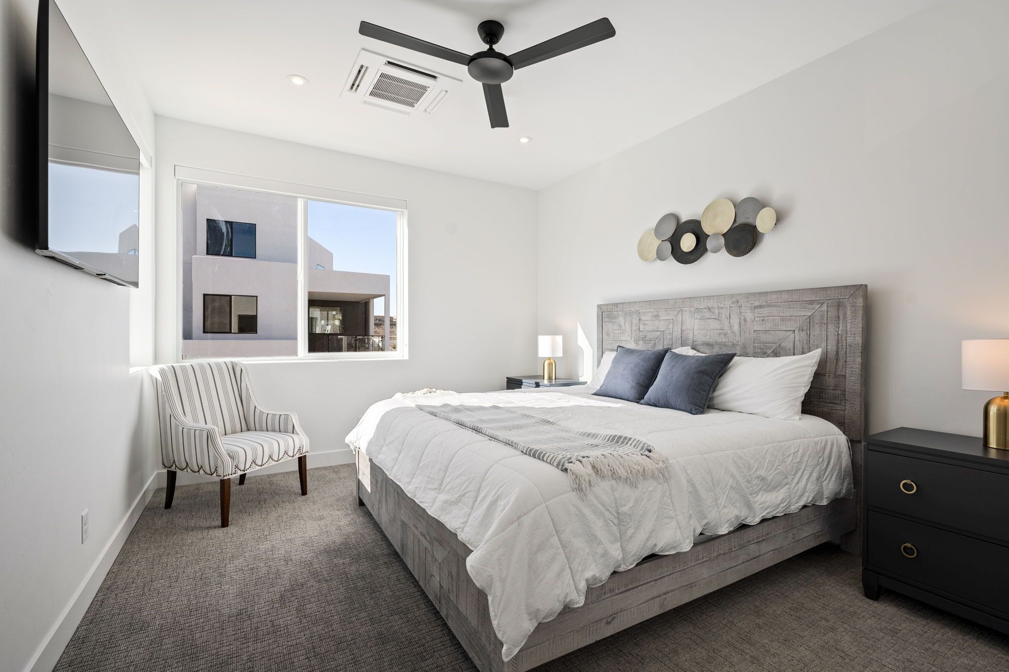 A bedroom with a bed, a chair, a nightstand, a wall-mounted lamp, and a ceiling fan.