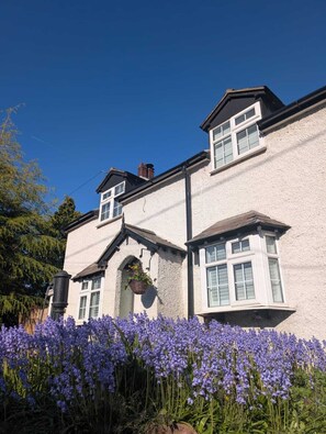 A two-story house with a white exterior, a visible chimney, and a flower bed with purple flowers in front. - Charming Cottage Near Chester Zoo + Cheshire Oaks (Mickle Trafford)