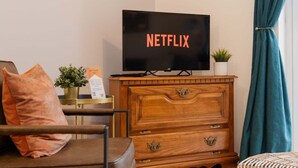 A room with a television displaying 'NETFLIX,' a wooden dresser, a chair, a small plant, and a window with curtains. - Stylish Chester Stay, Free Parking, Private Garden (Cheshire West and Chester)