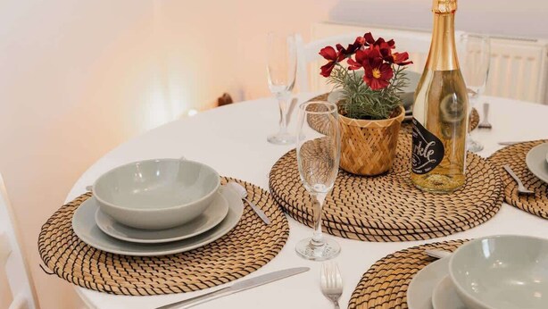 A round dining table with woven placemats, a bottle of sparkling wine, a small potted plant, and a bowl of red flowers. - Stylish Chester Stay, Free Parking, Private Garden (Cheshire West and Chester)