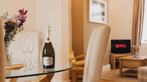 A living room with a glass table, a bottle of Prosecco, two wine glasses, a bouquet of flowers, a sofa, a TV displaying Netflix, and a framed picture on the wall. - Park and walk to city centre!  Cosy terrace (Handbridge)