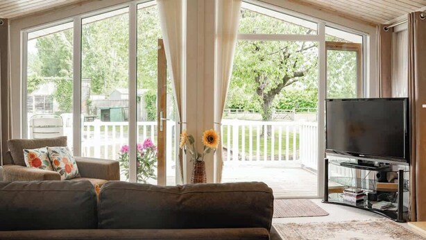 A living room with a sofa, a television, and a view of a backyard through large sliding glass doors. - Cosy Lodge on Farm with Donkeys, Parking (Chester)