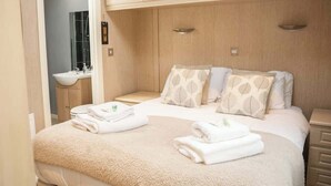 A hotel room with a bed, a nightstand, a bathroom with a sink, and wooden cabinets. - Cosy Lodge on Farm with Donkeys, Parking (Chester)