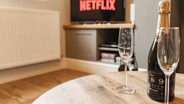 Quarto com uma mesa de madeira, uma garrafa de vinho e um copo. Uma televisão ao fundo exibe a palavra 'NETFLIX'.
