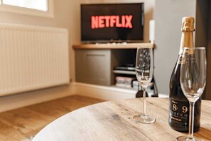 A room with a wooden table, a bottle of wine, and a glass. A television in the background displays the word 'NETFLIX'.