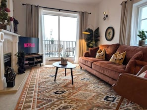 Una sala de estar con sofá, mesa de centro, chimenea, televisor y balcón con mobiliario exterior.