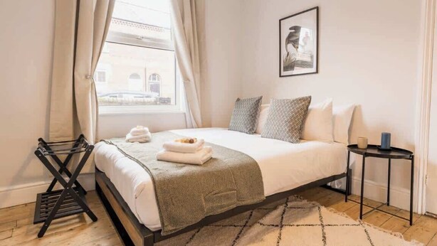 A hotel room with a bed, a window with curtains, a framed picture on the wall, and a small table with a lamp.