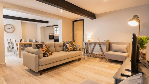 Ein modernes Wohnzimmer mit einer Couch, einem Sessel, einem Fernseher und einer Lampe. Im Hintergrund ist ein Essbereich zu sehen.