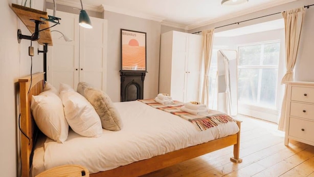 A bedroom with a wooden bed, a bedside lamp, a wall-mounted shelf, a framed picture, and a window with curtains. - Sleeps 6, off street parking, close to city (Cheshire West and Chester)