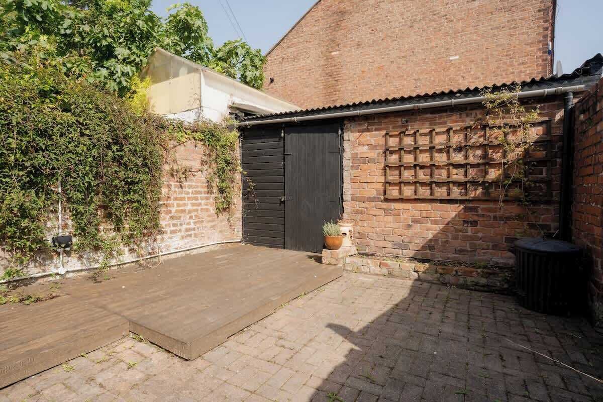 Un bâtiment en briques avec une porte fermée, un petit espace jardin avec une surface pavée et une plante en pot.