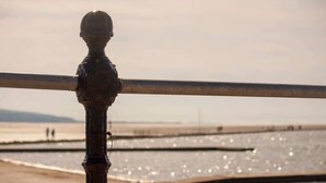 Une rambarde métallique agrémentée d’un élément décoratif, donnant sur une plage où la mer est calme et où l’on aperçoit des silhouettes au loin.