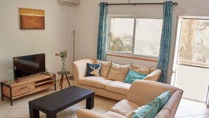 A living room with a sofa, a coffee table, a TV on a wooden stand, a vase with flowers, and a window with curtains.