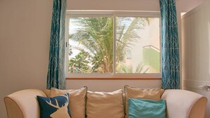 A beige sofa with four cushions against a wall, a window with blue curtains, and a view of palm trees outside.