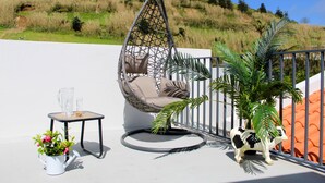 Una silla colgante con cojines, una mesita con una jarrón y vasos, una planta en maceta y una estatua decorativa de vaca en un balcón con vistas a una colina cubierta de hierba.