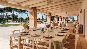 An outdoor dining area with a long table set for a meal, surrounded by chairs and a covered seating area with sofas and cushions. - Masseria Samali 14+4, Emma Villas (Leverano)