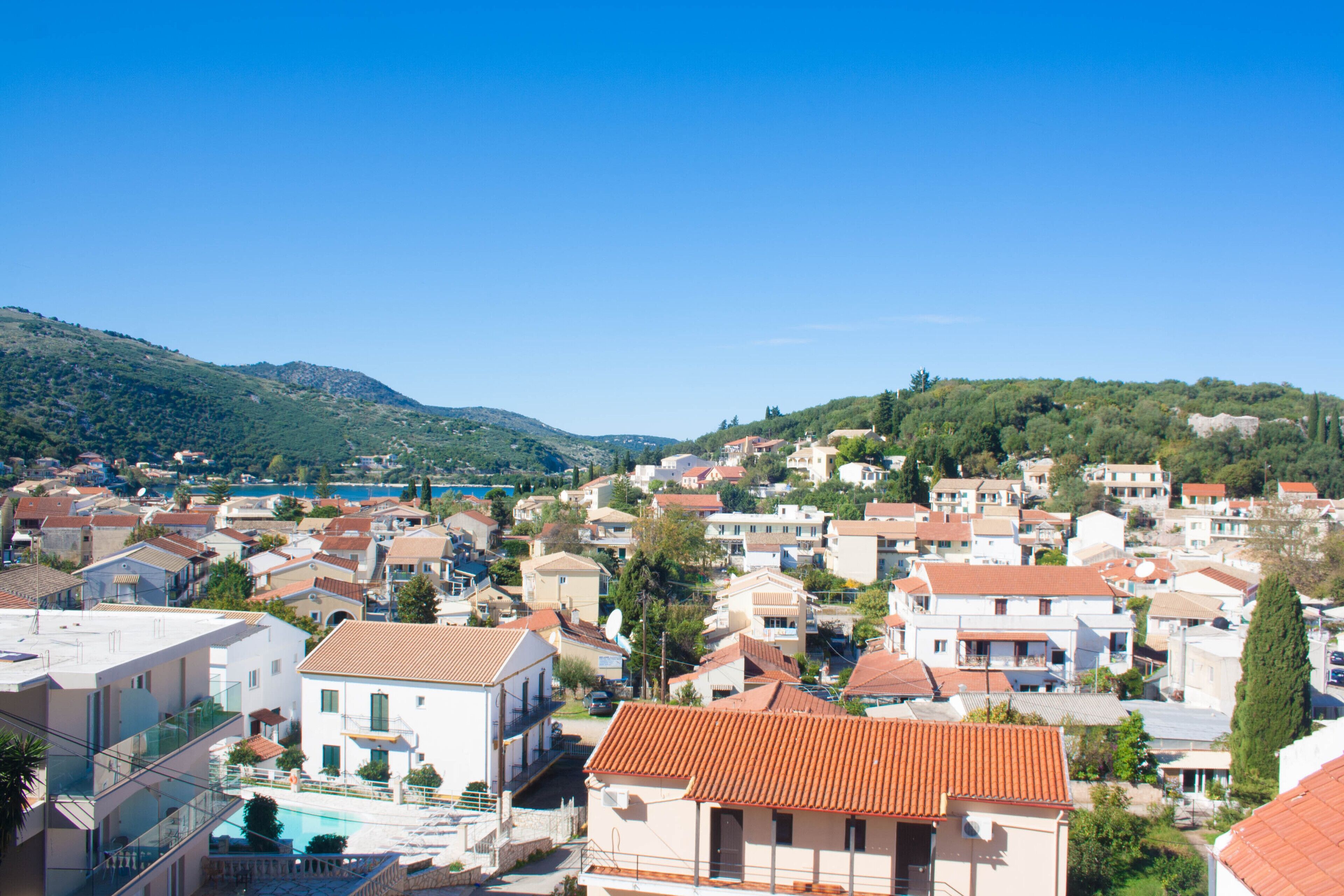 A coastal town with terracotta-roofed buildings, a clear blue sky, and a hilly backdrop.