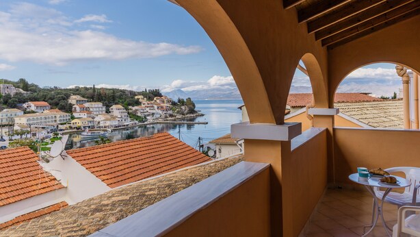 A balcony with a view of a coastal town, a table with a chair, and a building with red-tiled roofs.