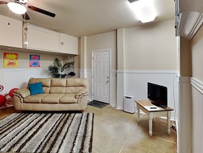 Een woonkamer met een beige bank, een gestreept vloerkleed, een televisie op een houten tafel en een plafondventilator.