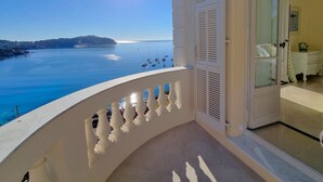 Un balcón con vista al mar y a barcos, una puerta blanca y una pequeña mesa con un florero.