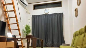 Una sala de estar con televisor, una mesa de madera, un sofá verde, una cortina oscura, un reloj y una ventana.