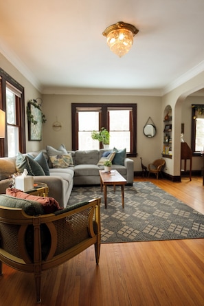 A living room with a sofa, a coffee table, a wicker chair, and a chandelier. - Charming Cottage with Hot Tub, Fire Pit, & Wooded Backyard (Saint Paul)