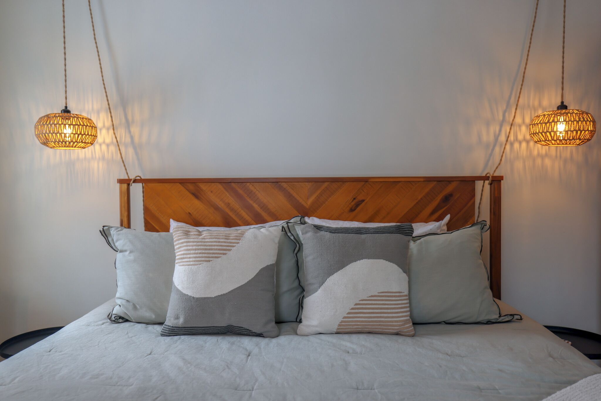 A neatly made bed with a wooden headboard, two hanging wicker lamps, and three pillows with abstract patterns.