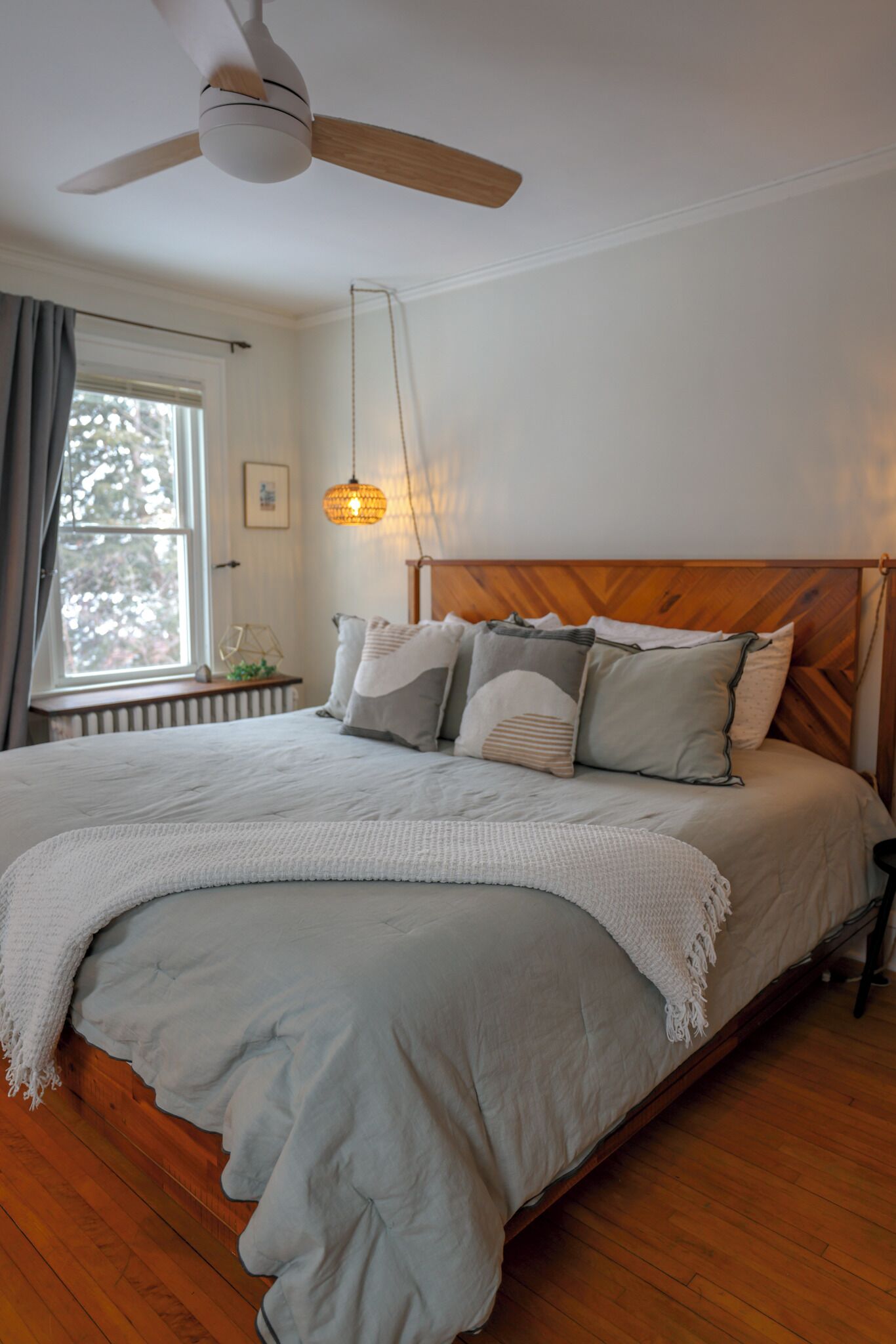 Ein Schlafzimmer mit einem Holz-Kopfteil, einem großen Bett mit vielen Kissen, einem Deckenventilator, einem Fenster mit Vorhängen und einer Pendelleuchte.