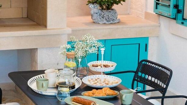 A cozy outdoor dining area with a table set for breakfast, including croissants and pastries, and a potted plant on a shelf.
