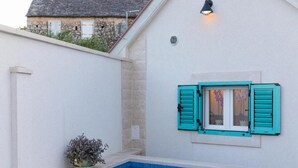 A small outdoor swimming pool with a tiled edge, a potted plant, and a window with turquoise shutters.