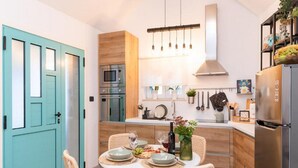 A modern kitchen with a dining area, featuring a teal door, wooden cabinets, stainless steel appliances, and a hanging light fixture.