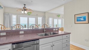 A kitchen with granite countertops, a stainless steel dishwasher, and a view of the ocean through large windows. - Oceanfront Condo w/ Private Deck, Resort Pool, Hot Tub, Clubhouse, Beach Access (Hatteras)