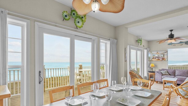 A dining area with a view of the ocean, a balcony with a wooden railing, and a living area with a sofa and a television. - Oceanfront Condo w/ Private Deck, Resort Pool, Hot Tub, Clubhouse, Beach Access (Hatteras)