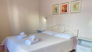 Ein ordentlich bezogenes Bett mit weißer, gesteppter Bettdecke, ein Nachttisch mit Lampe sowie zwei gerahmte Kunstwerke an der Wand.