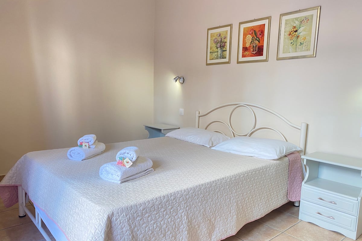 Un lit bien fait, recouvert d’une courtepointe blanche matelassée, une table de chevet avec une lampe, et deux œuvres d’art encadrées accrochées au mur.