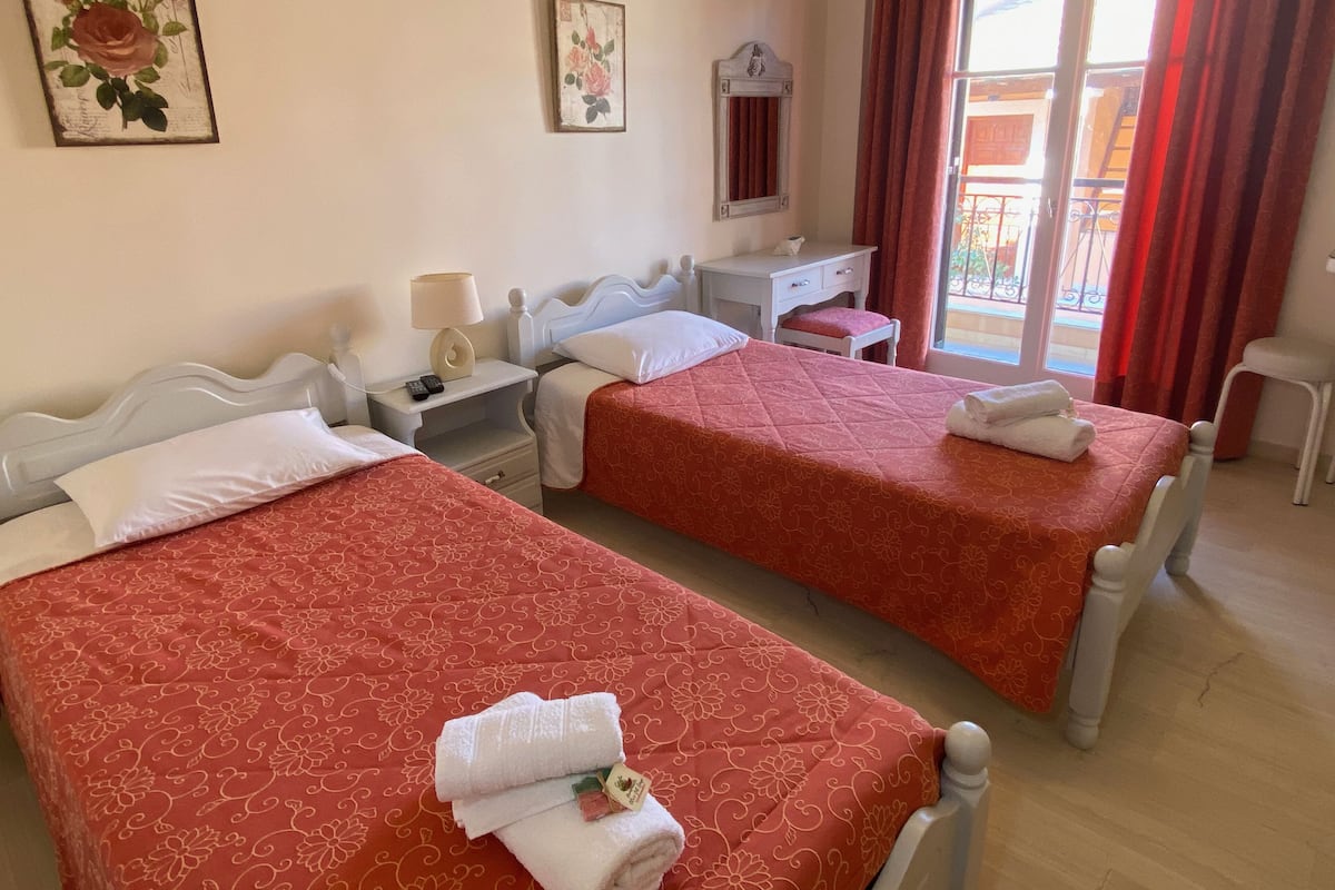 Une chambre d’hôtel avec deux lits, un bureau, une chaise et un balcon avec des rideaux rouges.