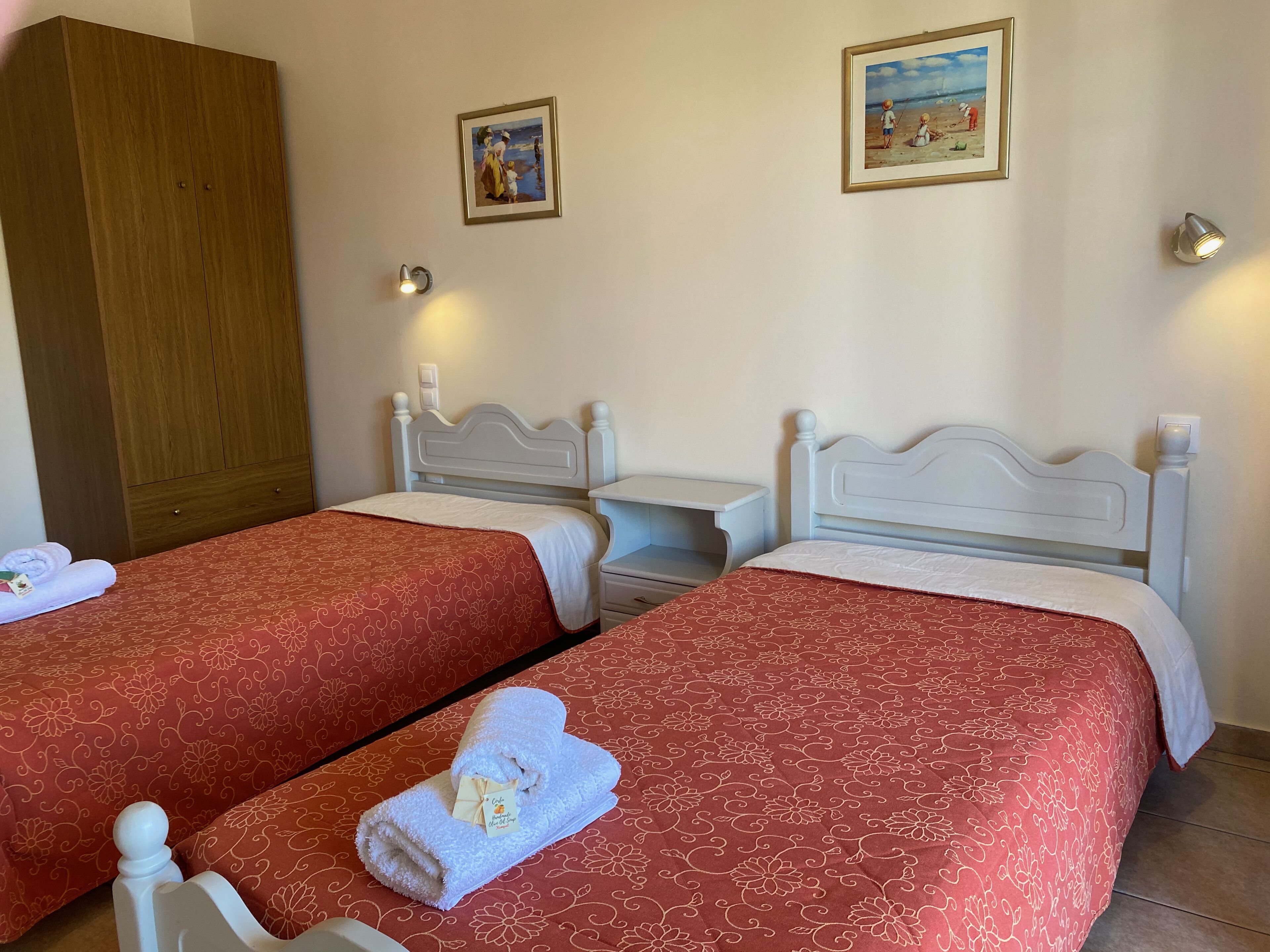 A hotel room with two single beds, each with a red floral bedspread and white headboards. There are two wall-mounted light fixtures, a wooden wardrobe, and two framed pictures on the walls.